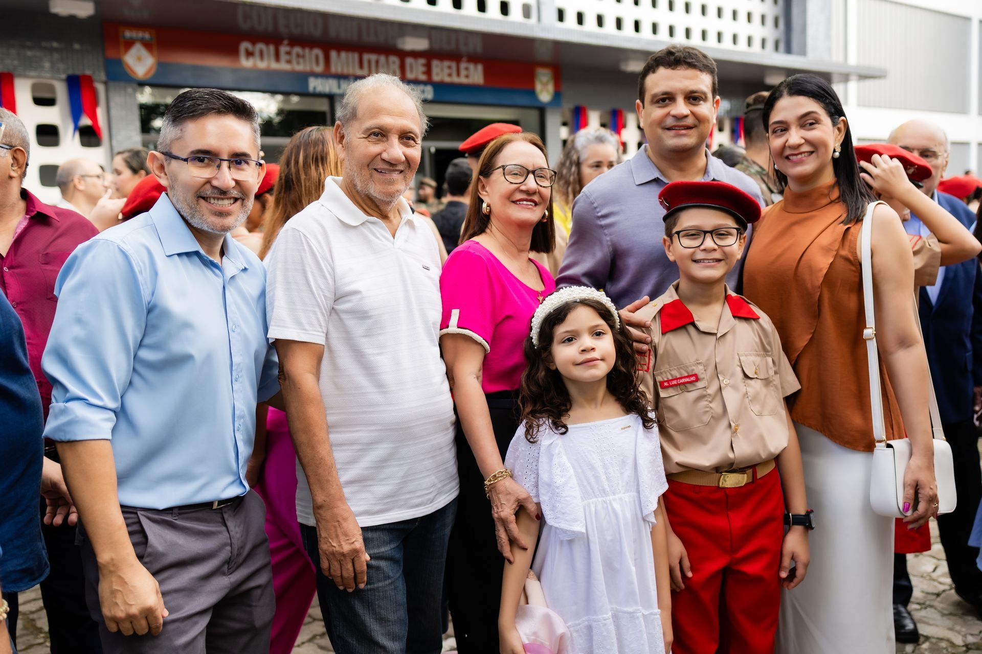 Grupo de pessoas sorrindo em frente a um prédio. Uma criança de uniforme e uma mulher de branco estão em primeiro plano.