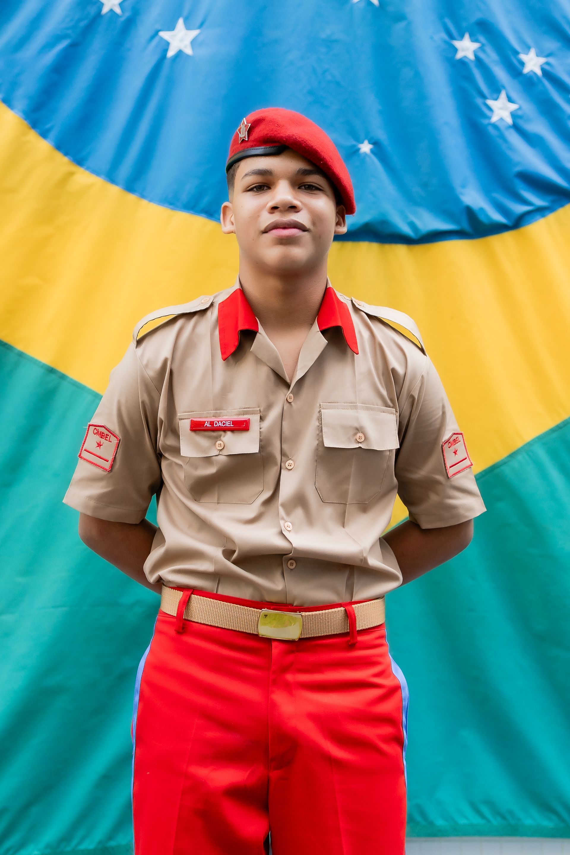 Um jovem de boina vermelha e uniforme está em frente a uma bandeira brasileira.
