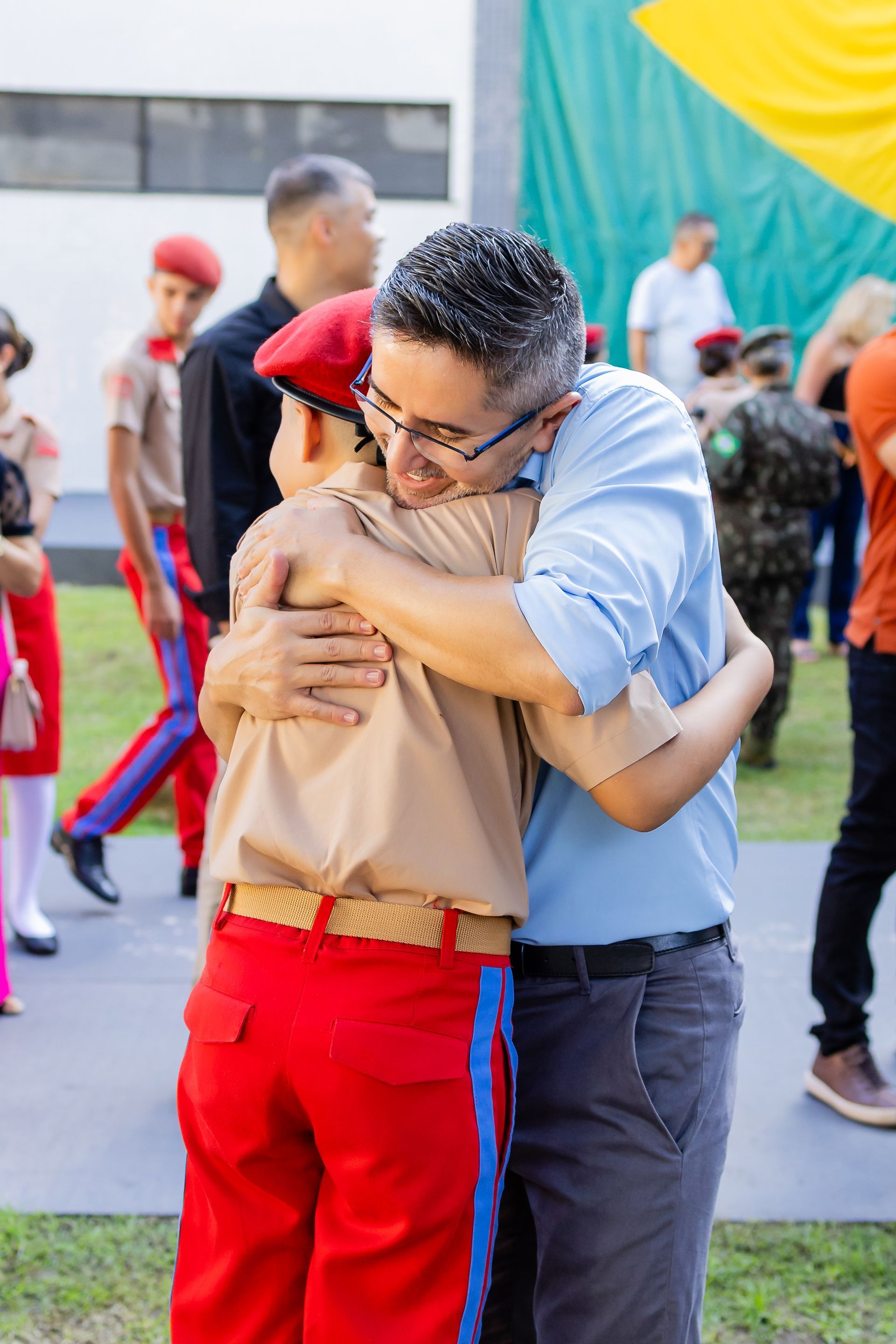 Homem abraçando uma criança uniformizada, do lado de fora. Menino de calça vermelha, camisa bege e boina vermelha; homem de camisa azul e calça cinza.