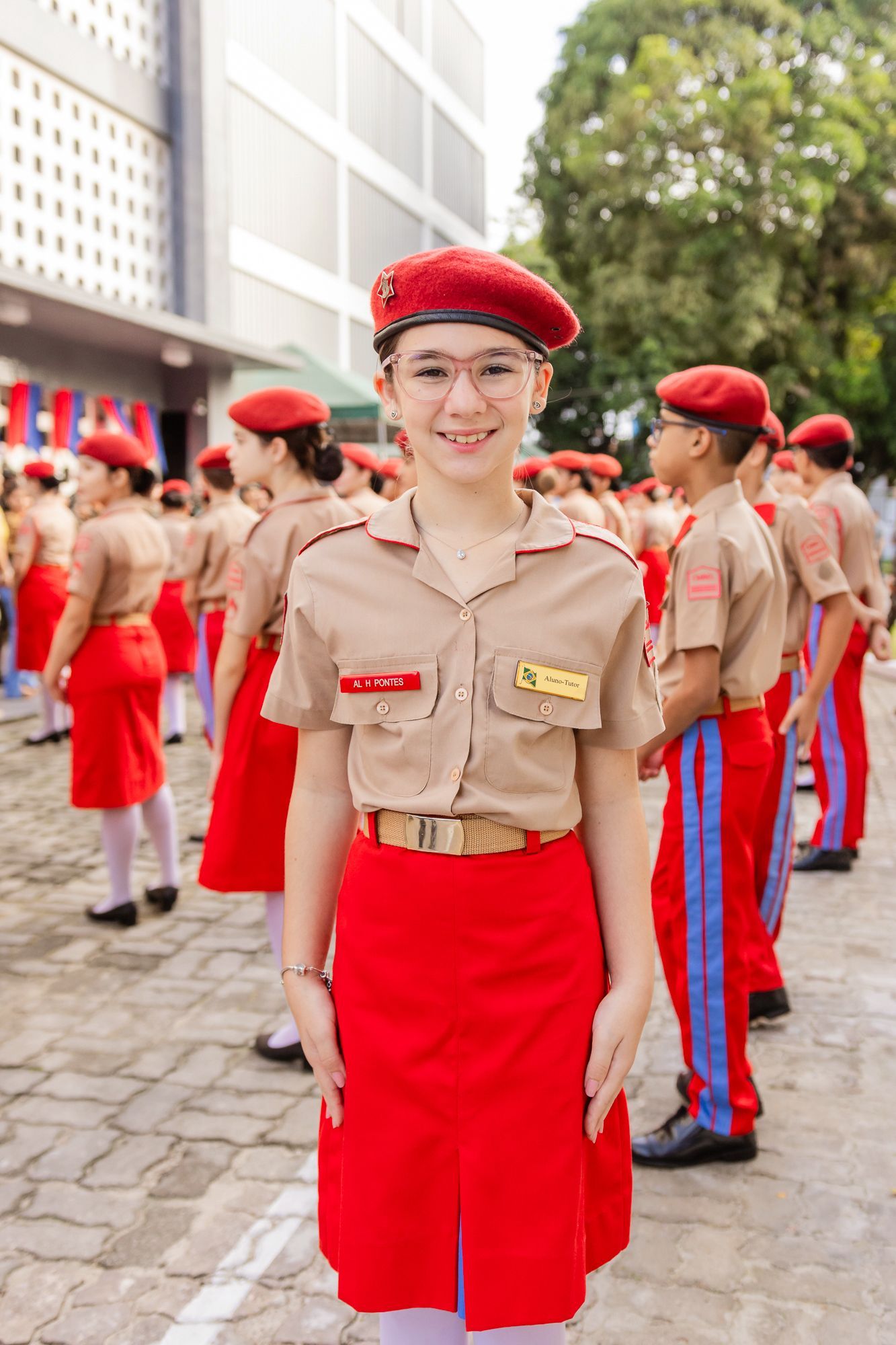 Garota de uniforme vermelho e cáqui sorri em formação com outras pessoas.