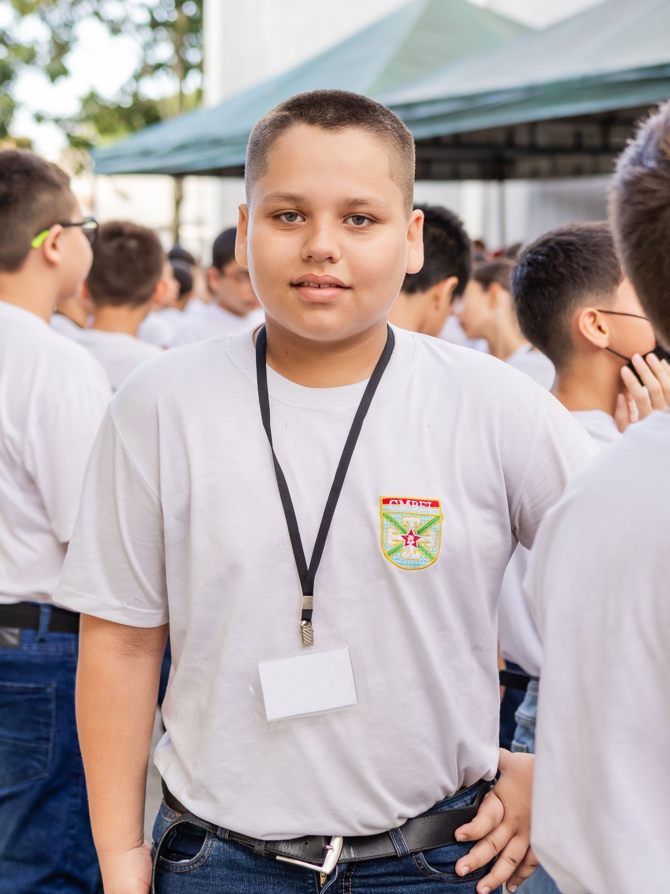 Menino de camisa branca com identidade, olhando para a câmera, em pé do lado de fora com outras pessoas.
