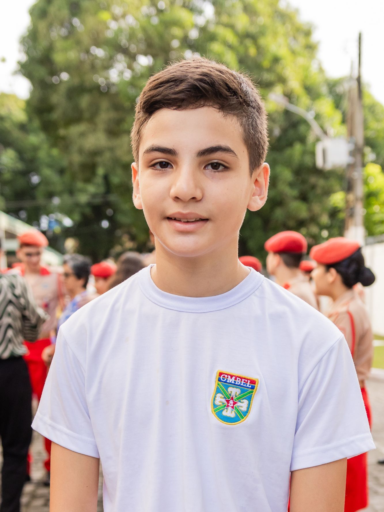 Menino de camisa branca sorri, em pé ao ar livre, perto de pessoas com chapéus vermelhos.