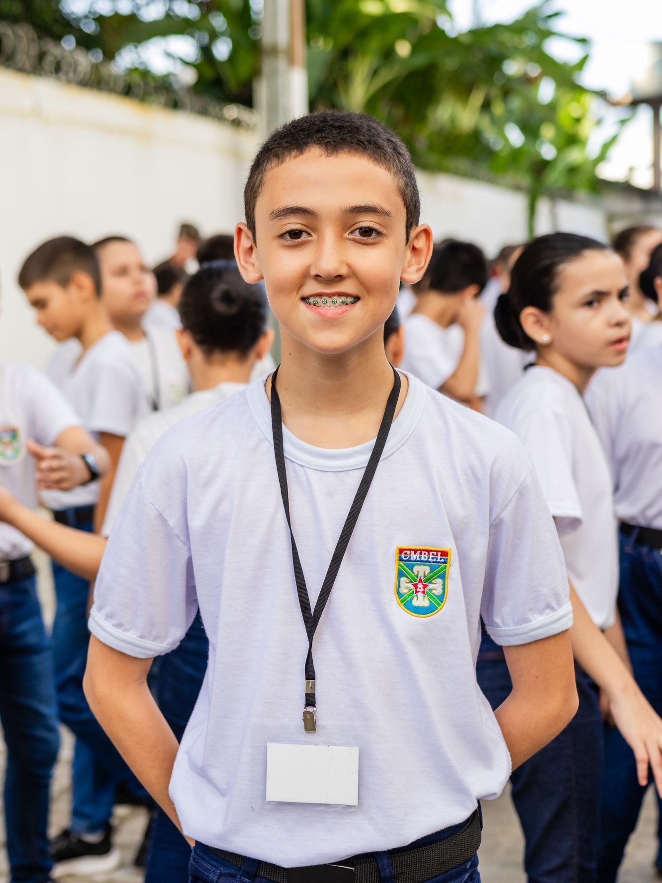 Menino com suspensórios e cordão, sorri, em pé na frente de um grupo vestindo camisas brancas e calças escuras.