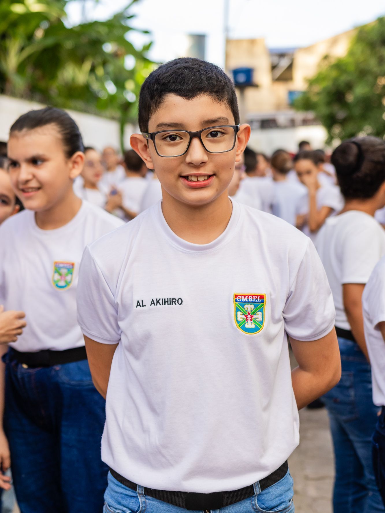 Menino de óculos, vestindo uma camisa branca com o emblema da escola, sorrindo, em pé do lado de fora com outros alunos.