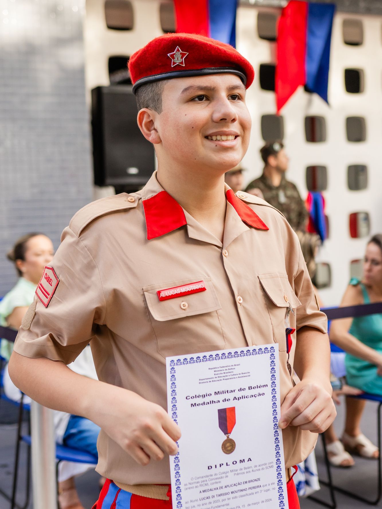Adolescente de uniforme bege e boina vermelha segura certificado, sorrindo. Evento ao ar livre.