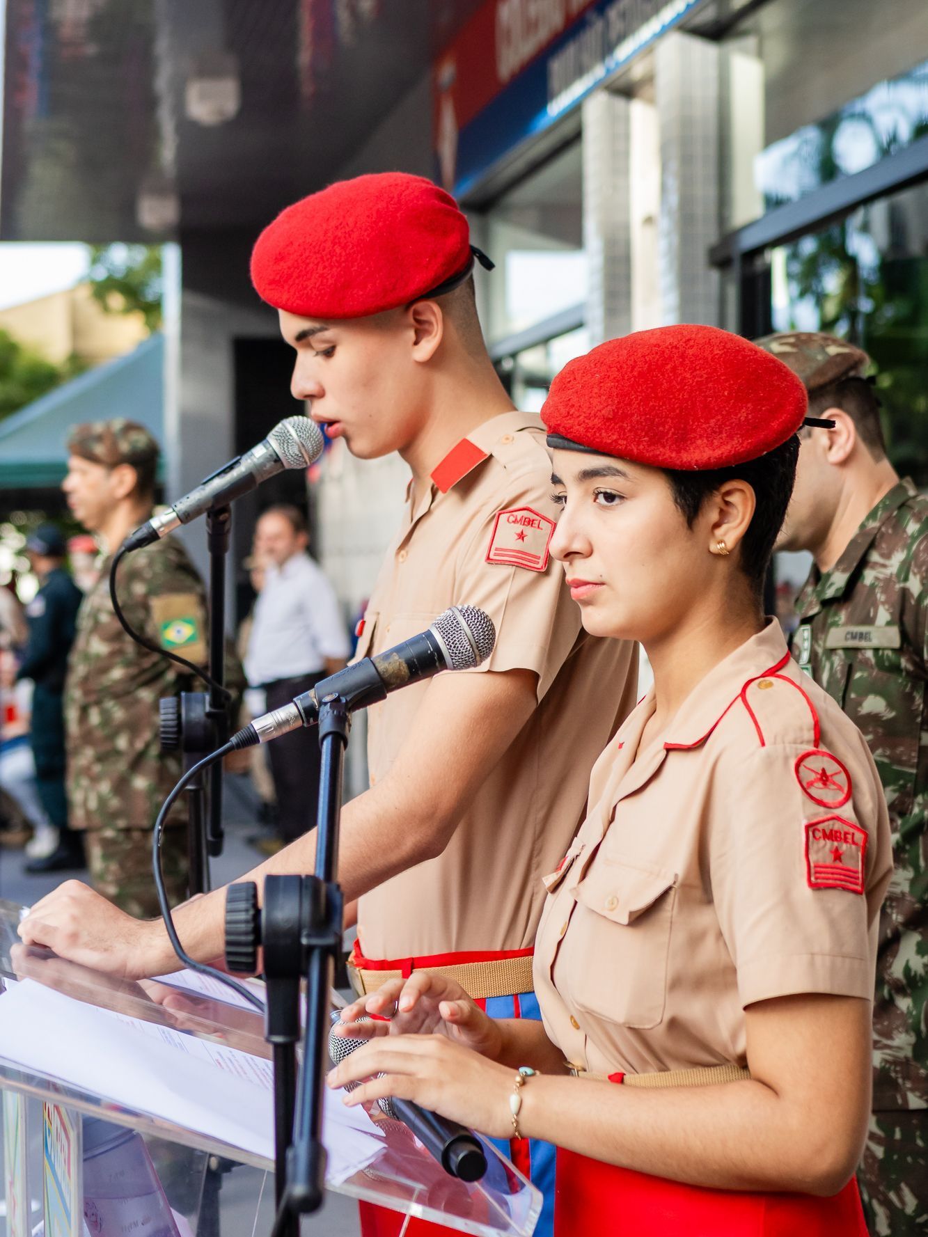 Duas pessoas de boinas vermelhas e uniformes bege falam ao microfone. Ambiente externo.