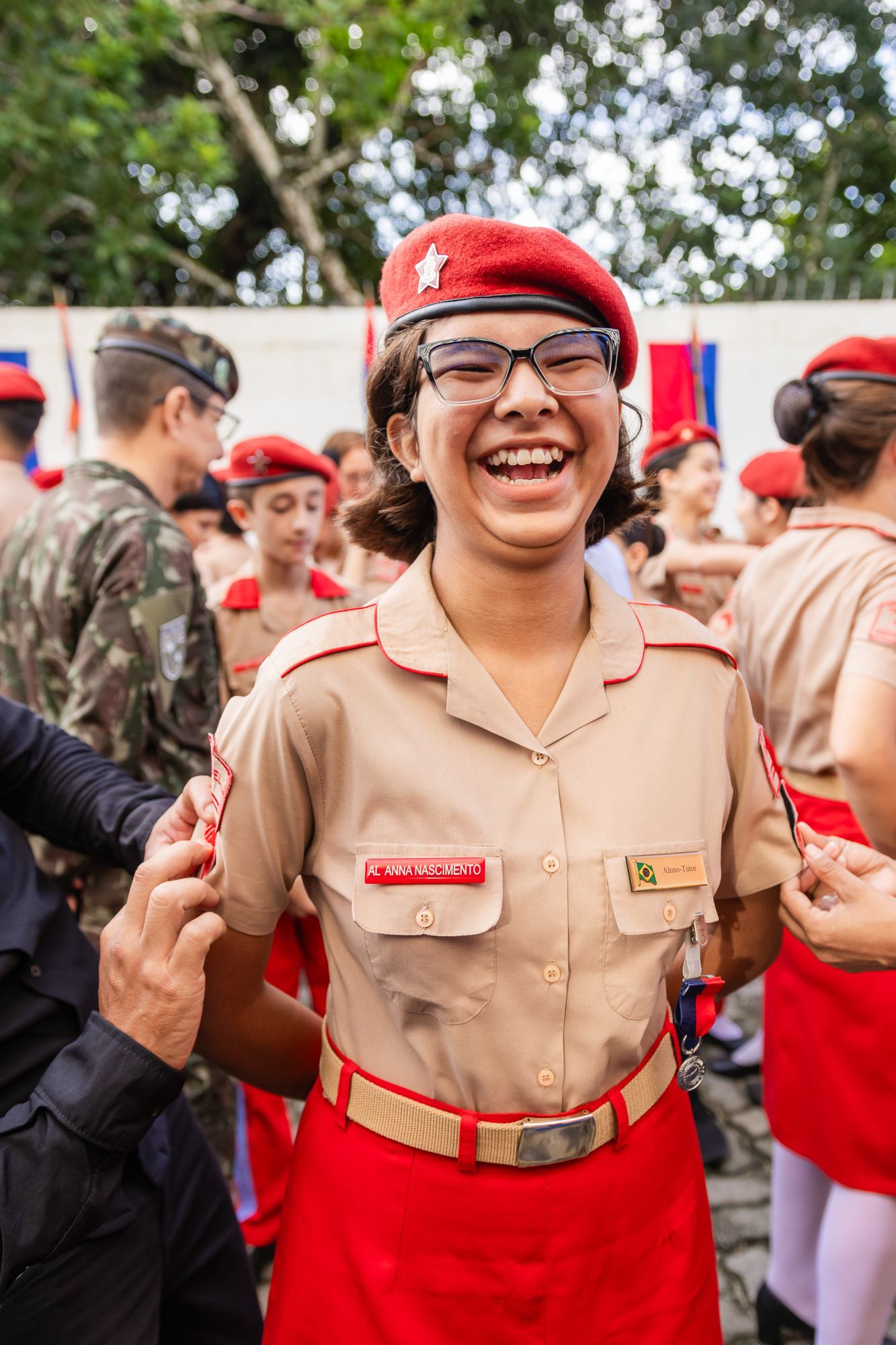 Garota de uniforme, boina vermelha, sorri alegremente. Rodeada por outras pessoas com trajes semelhantes, ao ar livre.