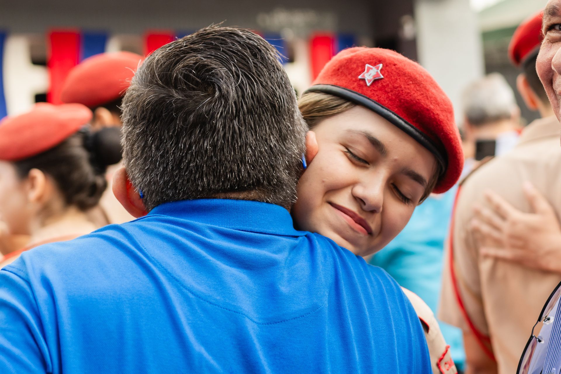 Mulher de boina vermelha abraça um homem de camisa azul, sorrindo e de olhos fechados. Evento ao ar livre.