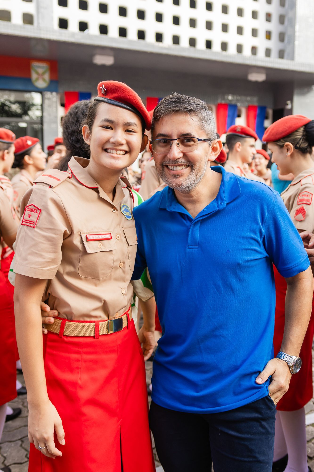 Uma jovem uniformizada e um homem de camisa azul sorriem, posando ao ar livre com outros uniformizados.