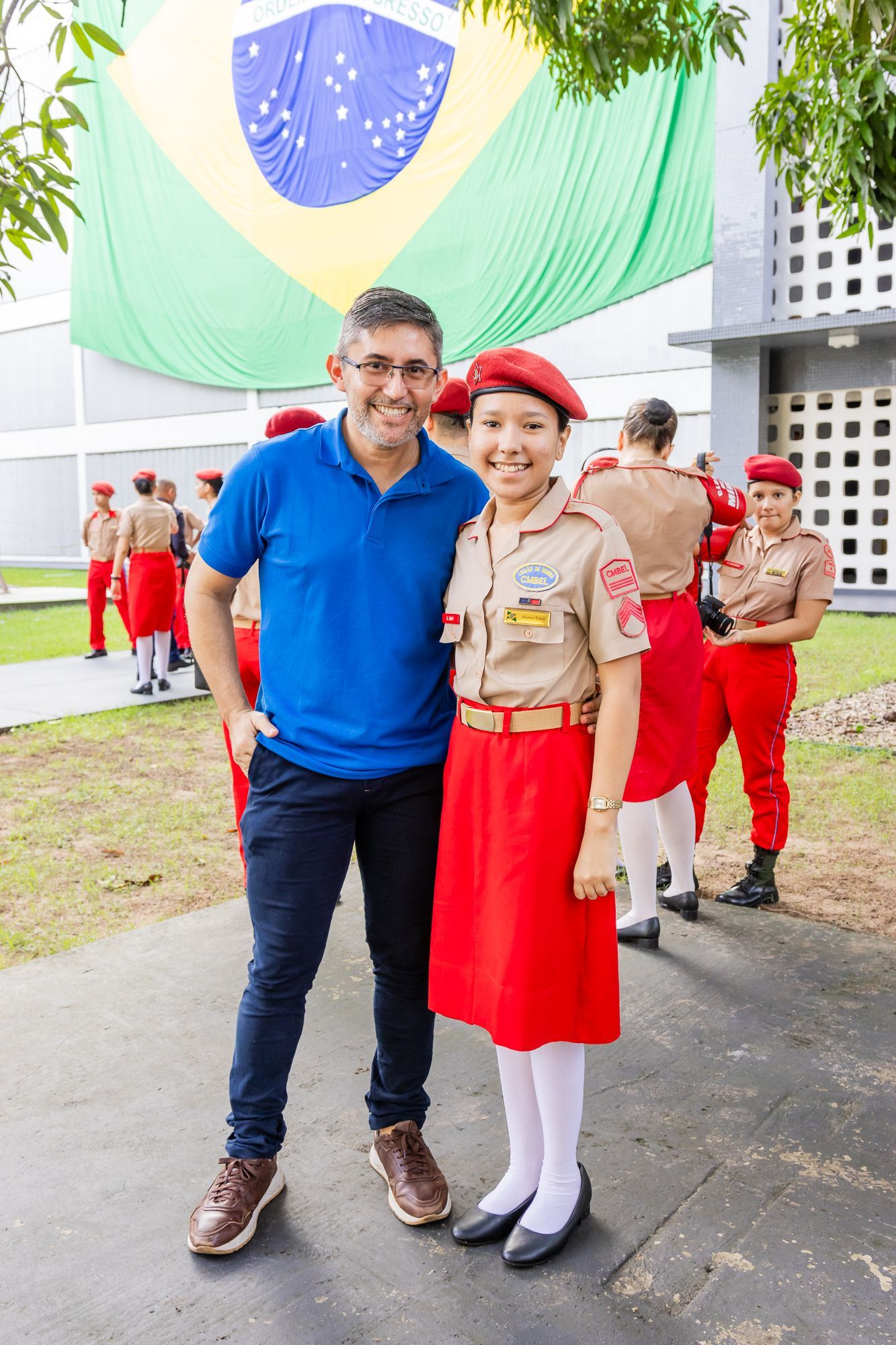 Homem e jovem uniformizados posam em frente a uma grande bandeira brasileira; outras pessoas uniformizadas estão ao fundo.