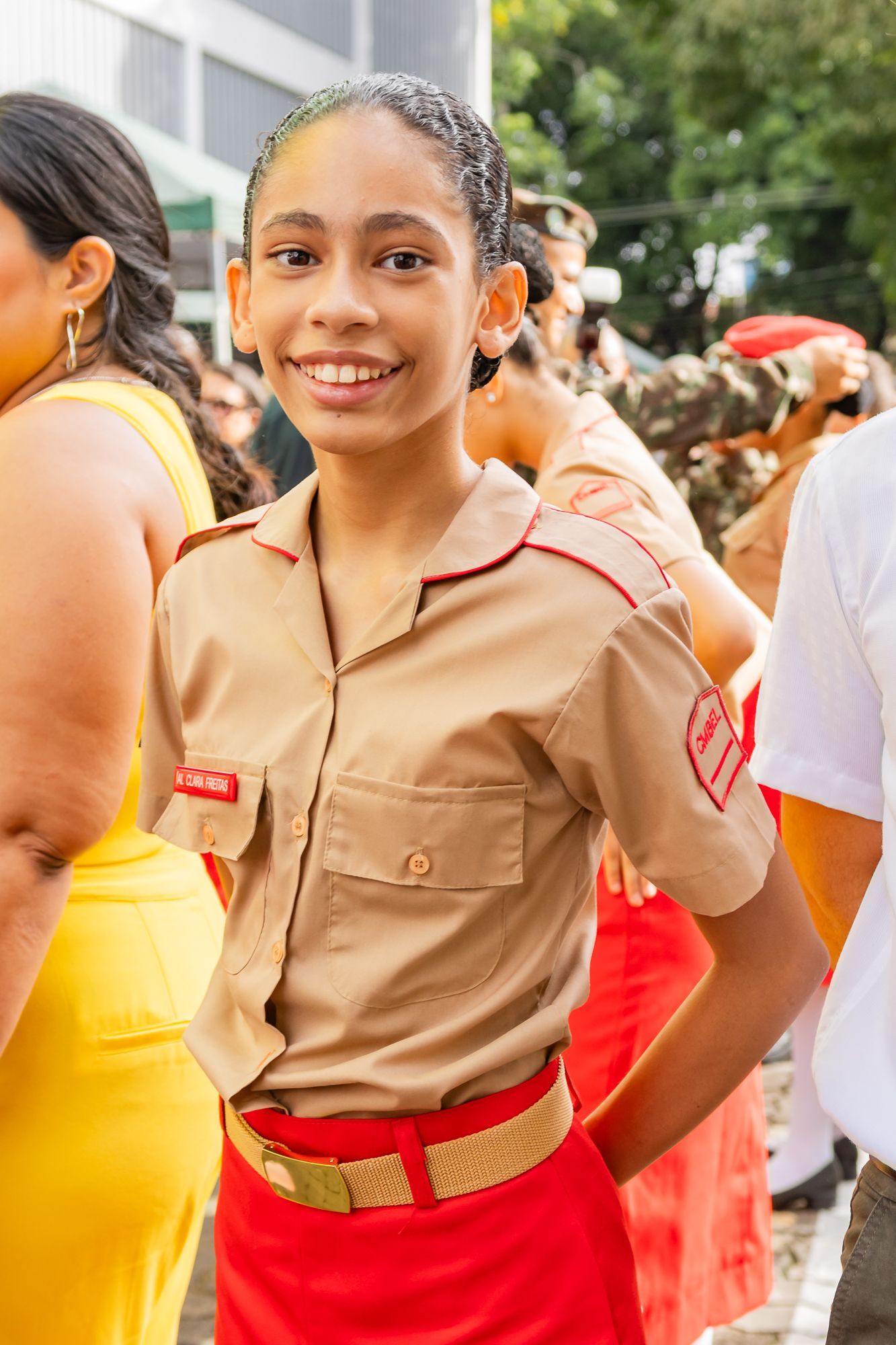 Jovem de camisa bege e saia vermelha, sorrindo, ao ar livre.