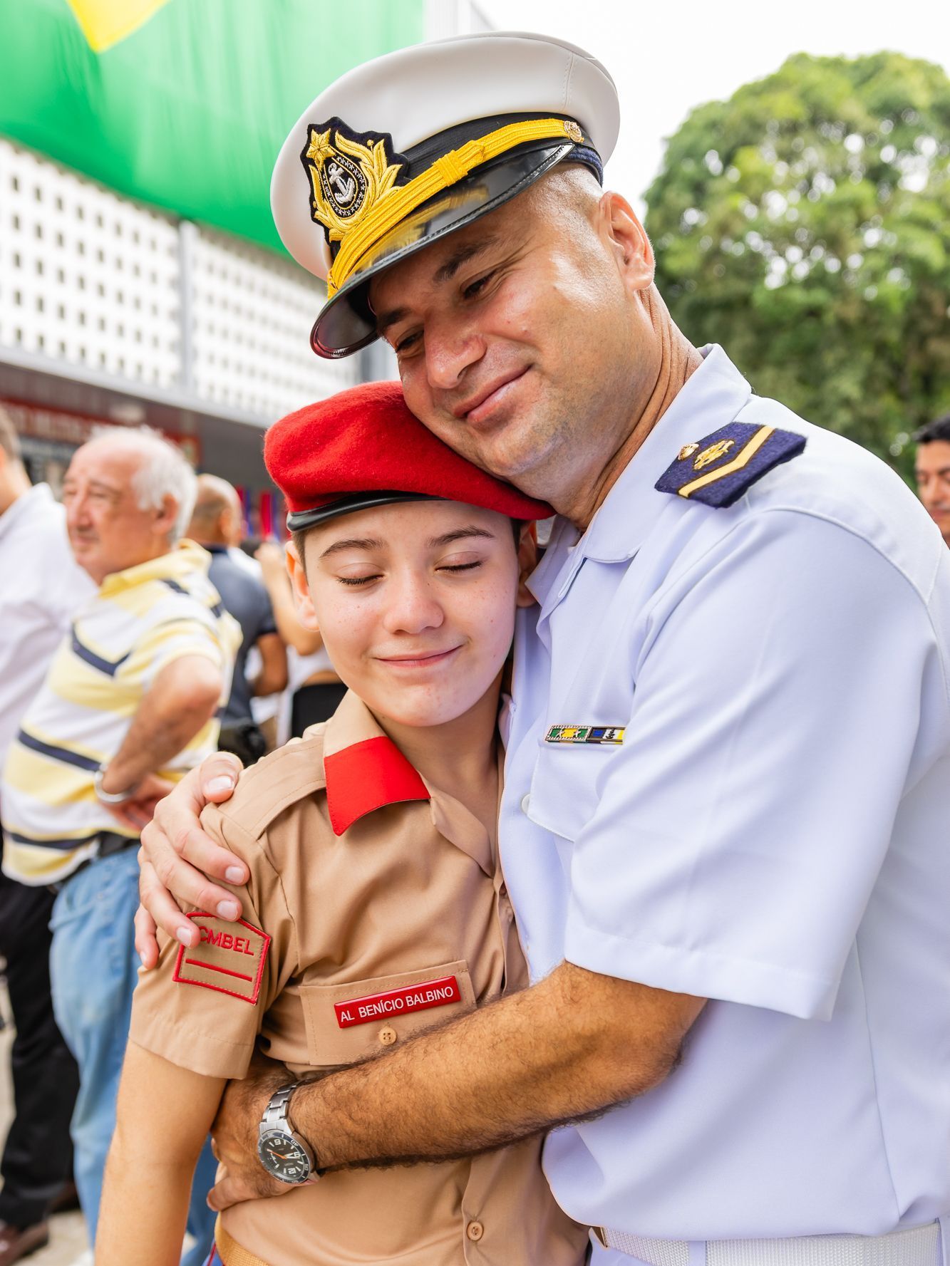 Homem em uniforme naval abraça criança em uniforme de cadete; ambos sorrindo. Ao ar livre, ensolarado.