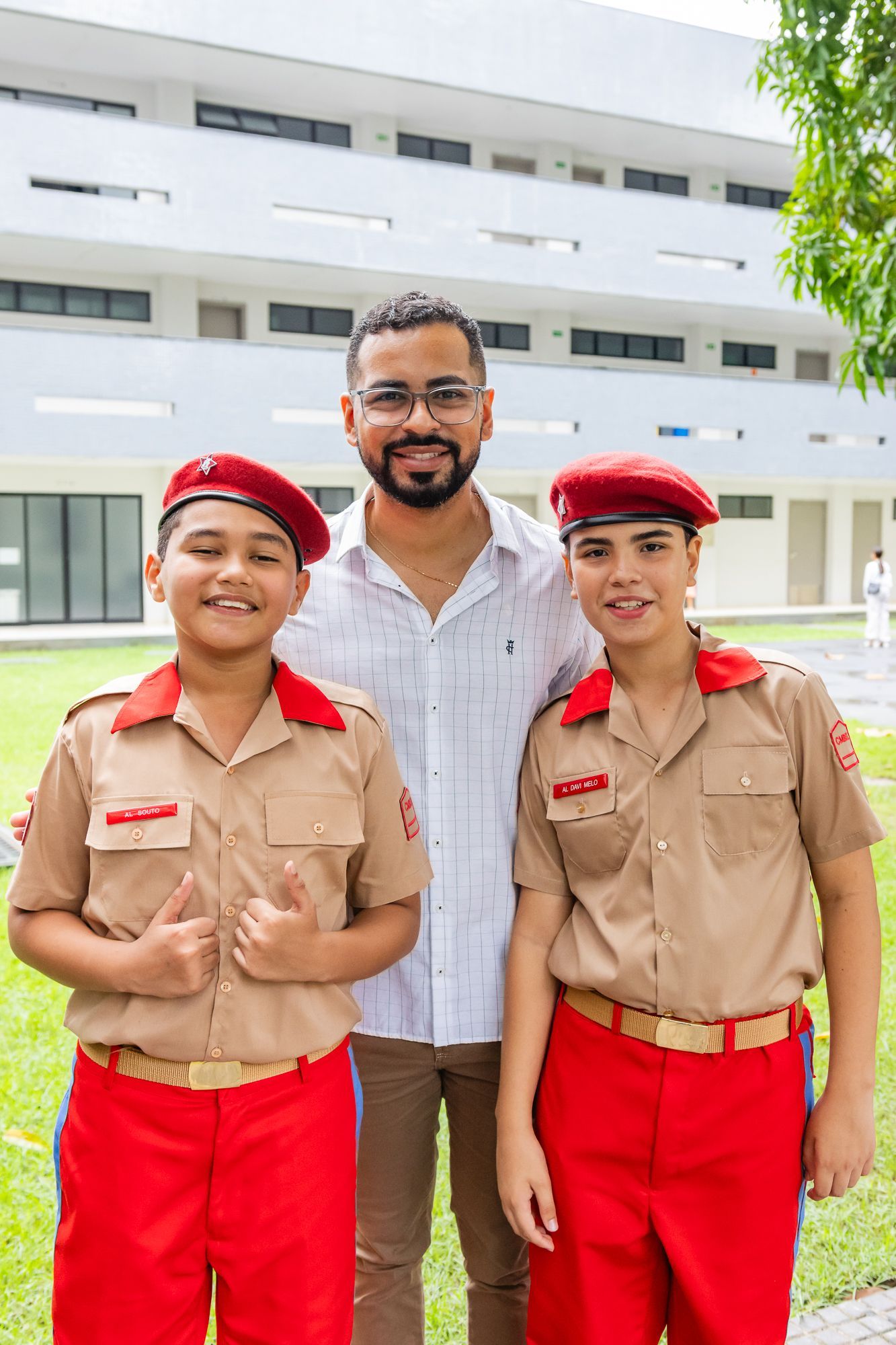 Três pessoas posando em frente a um edifício moderno; dois meninos em uniformes vermelhos/bege e um homem sorrindo.