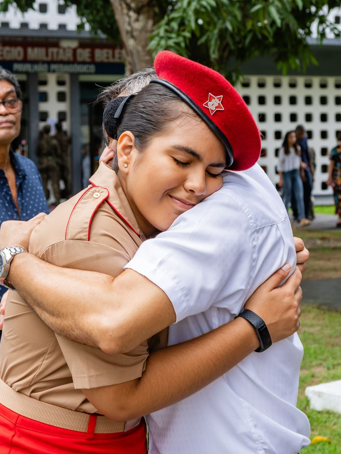 Mulher de uniforme militar abraça alguém, sorrindo, ao ar livre. Boina vermelha, camisa marrom-clara.