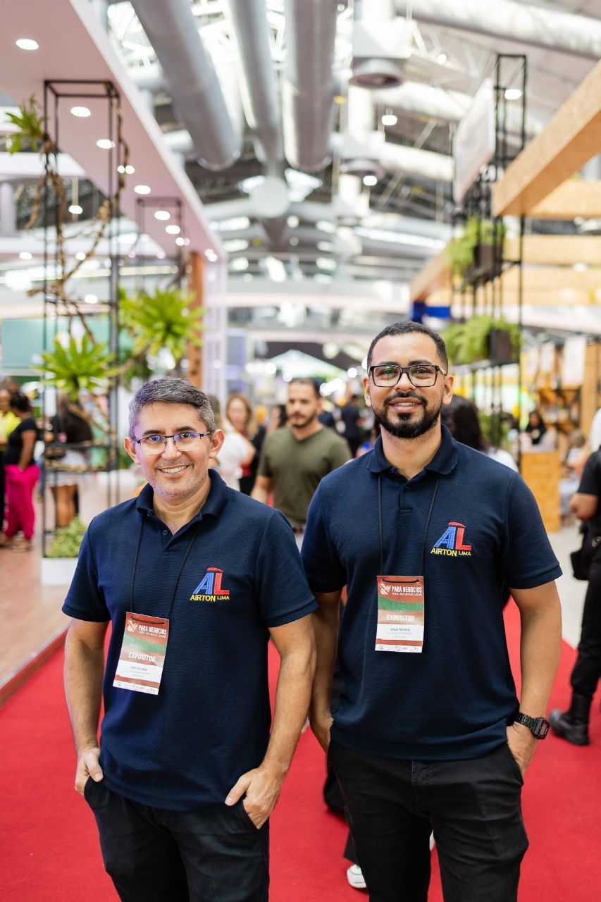 Dois homens vestindo camisas azul-marinho com logotipo, sorrindo, posam em um tapete vermelho em uma feira comercial.
