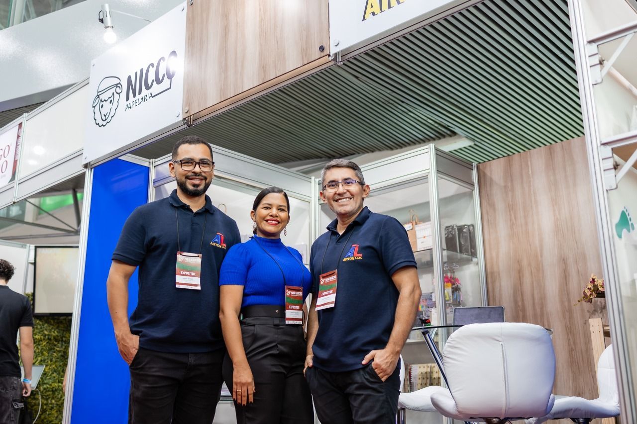 Três pessoas sorrindo em um estande em um salão de convenções; dois homens de camisa azul-marinho e uma mulher de azul.