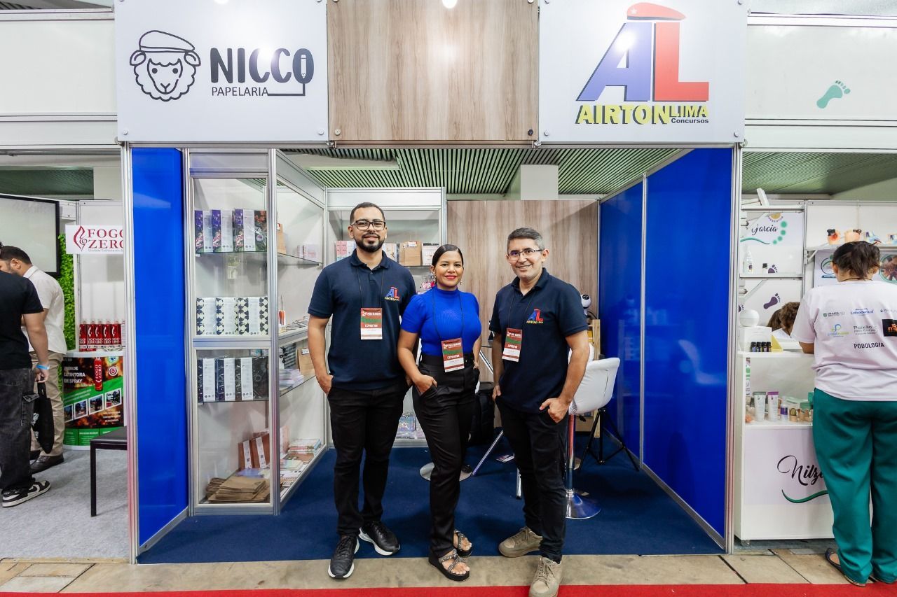 Três pessoas estão em frente a um estande de uma feira comercial com logotipos e produtos expostos, sorrindo para a câmera.