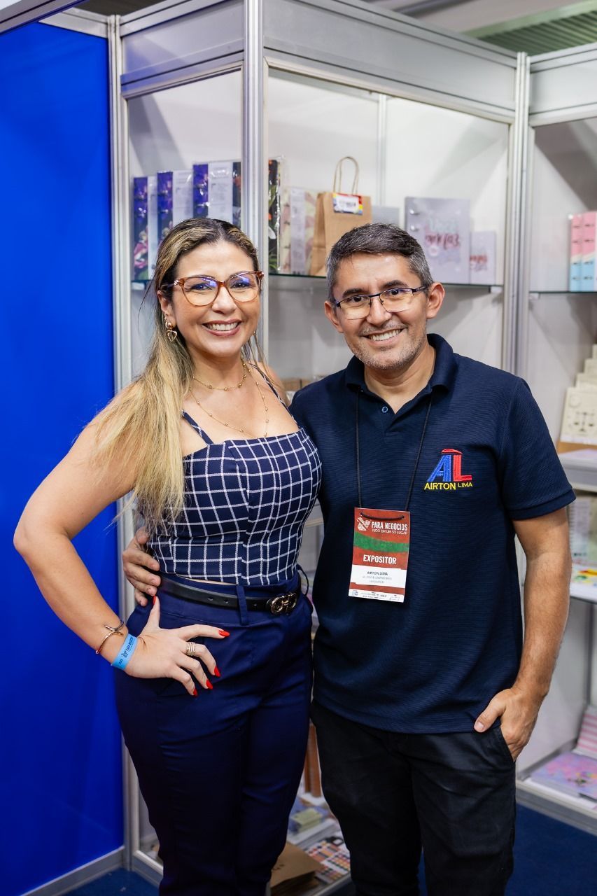 Mulher e homem sorrindo, posando em uma cabine. A mulher usa óculos e uma roupa azul. O homem veste uma camisa azul-marinho.