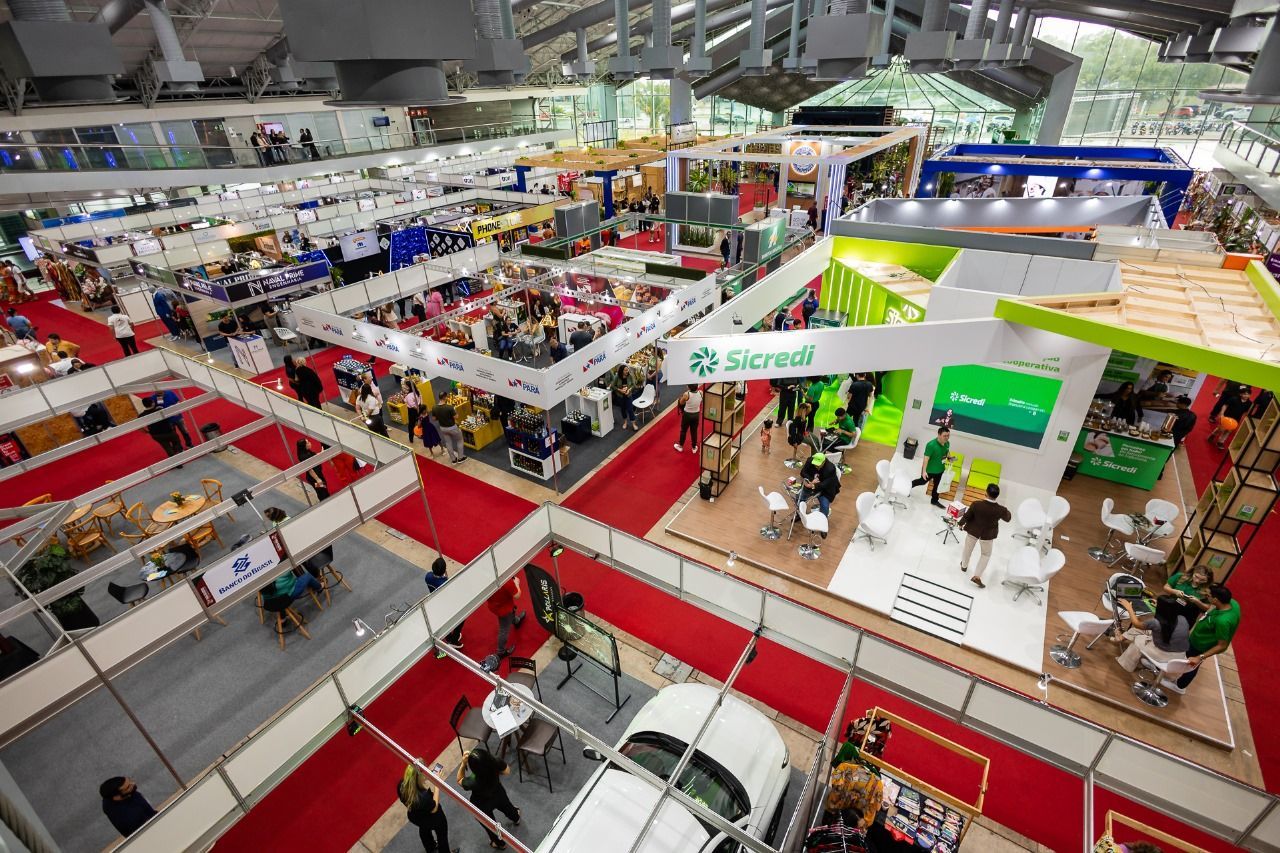 Vista de cima de uma feira com estandes e displays dentro de um grande salão. Tapetes vermelhos e muitos participantes são visíveis.