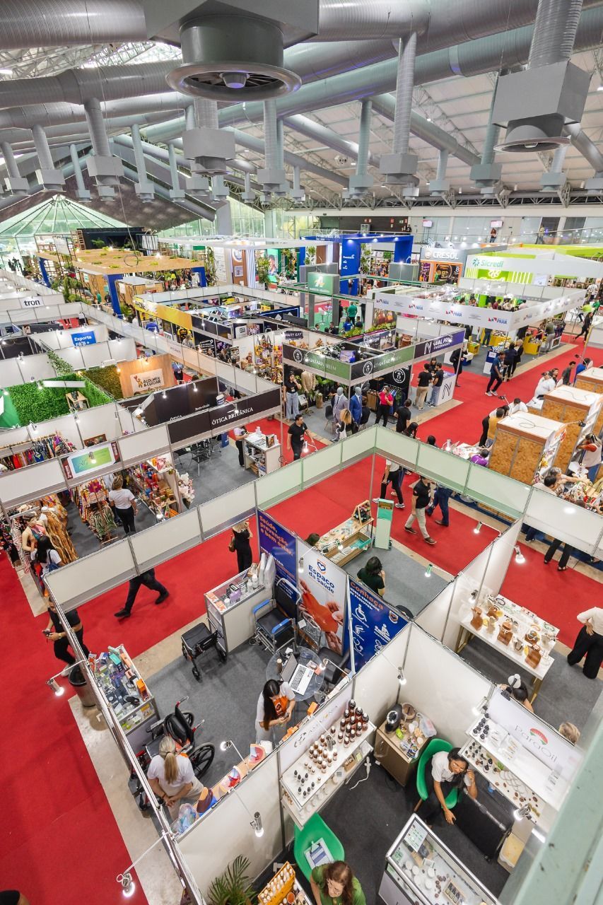 Vista aérea de uma grande feira comercial com estandes e pessoas circulando. Tapete vermelho e teto branco.