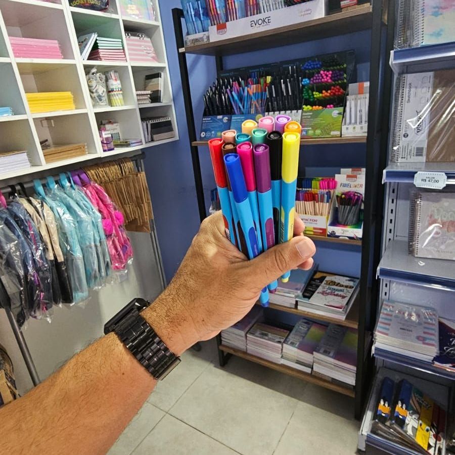 Mão segurando marcadores coloridos em uma papelaria, prateleiras com vários suprimentos ao fundo.