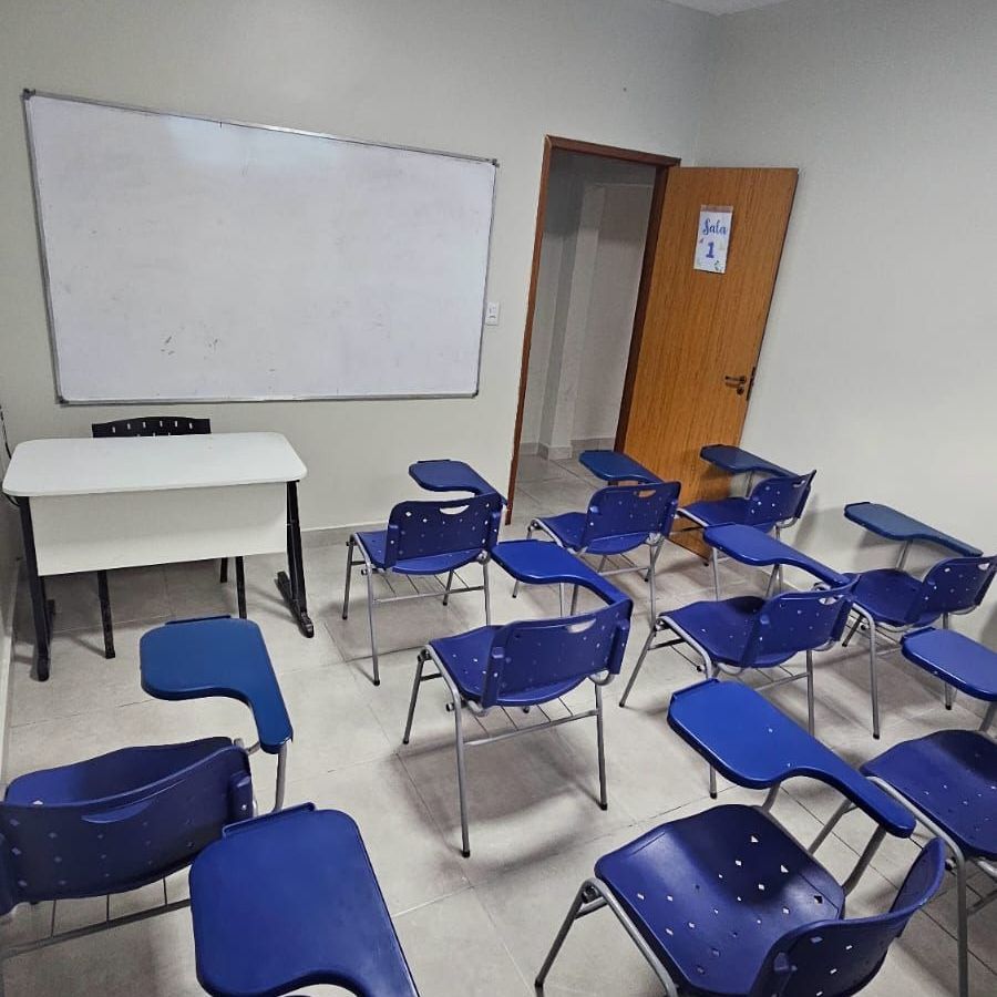 Sala de aula com cadeiras azuis, quadro branco, mesa e porta aberta.