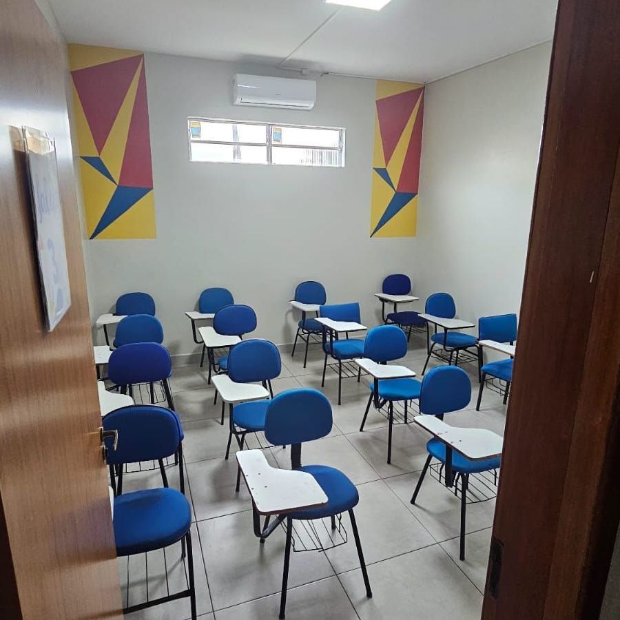 Sala de aula com cadeiras azuis, carteiras brancas e arte geométrica na parede.
