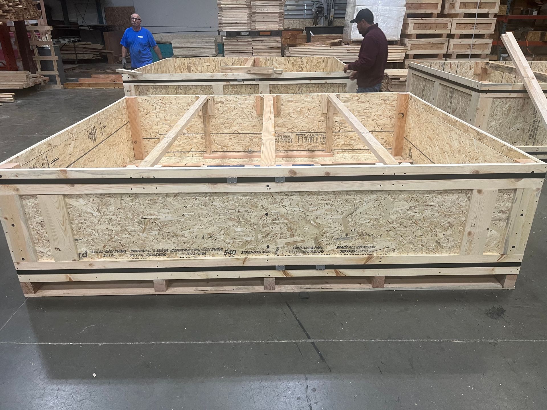 Large wooden shipping crate, possibly for machinery, in a warehouse setting, with lifting straps.