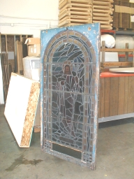 Stained glass window depicting a figure, set in a blue wooden frame, leans indoors against a wall.