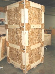 Large, wooden shipping crate made from plywood and corner supports, sitting on a pallet in a warehouse.
