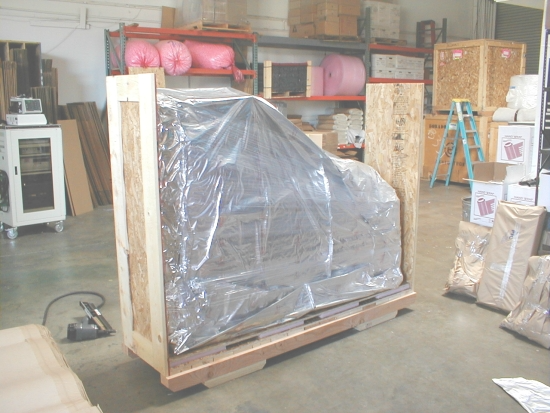 Piano wrapped in plastic and secured in a wooden crate, in a warehouse setting.