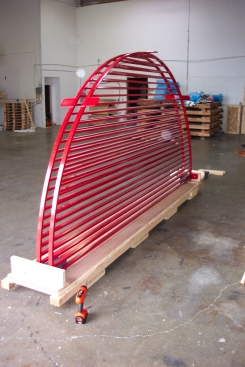Red arched metal sculpture with horizontal slats, resting on wooden supports in a warehouse.