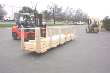 Two forklifts lifting and transporting a large wooden crate in a parking lot.