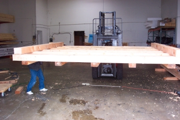 A forklift carrying a wooden frame in a warehouse, with a person walking beside it.