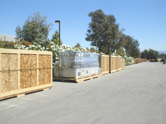 Wooden crates and a metal-wrapped object sit on pallets along a paved area, trees and bushes behind.