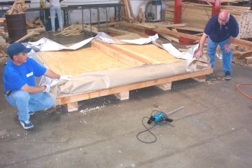 Two workers wrapping large wooden panels on a pallet in a warehouse. A power drill is on the floor.