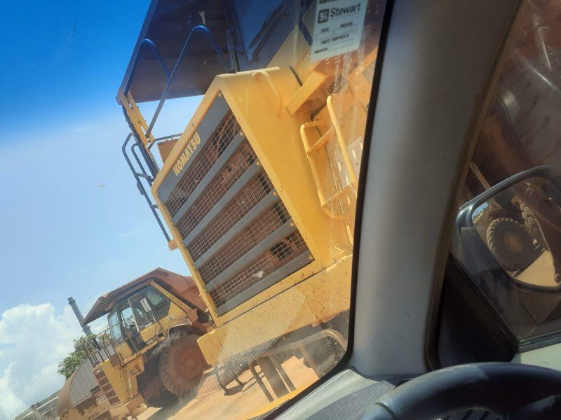 Yellow construction equipment, likely a grader, seen through a windshield on a sunny day.
