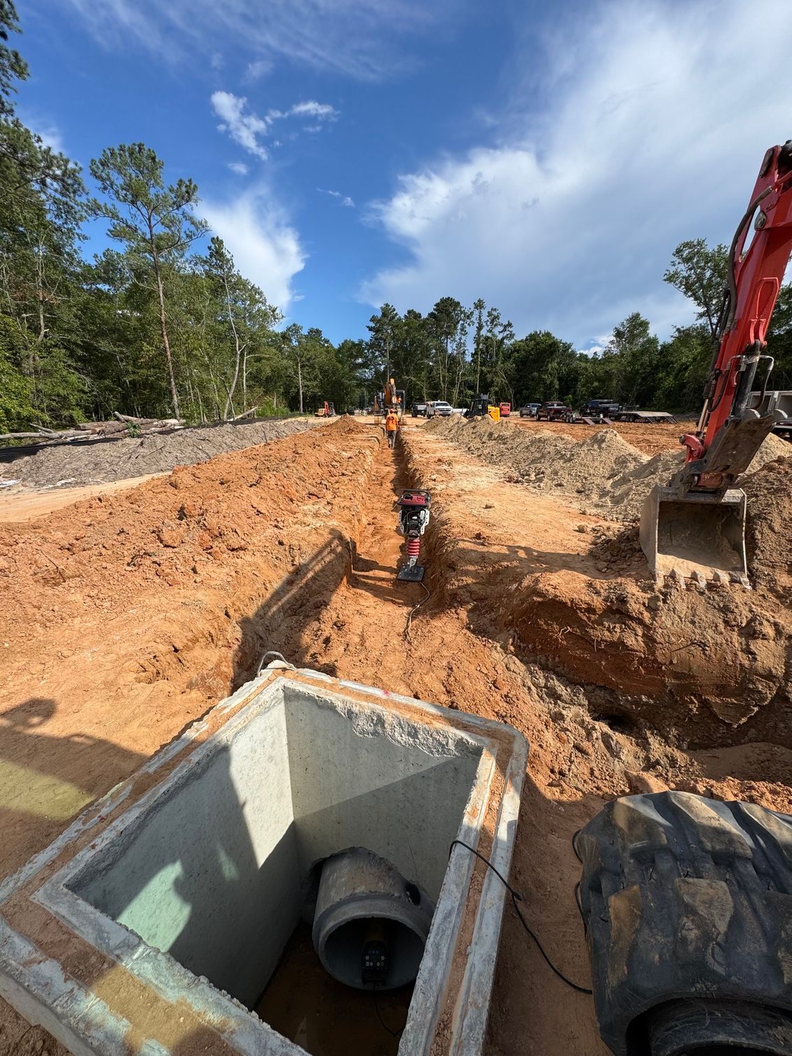 Construction trench with exposed pipes, workers, and excavator in a dirt excavation site under a blue sky