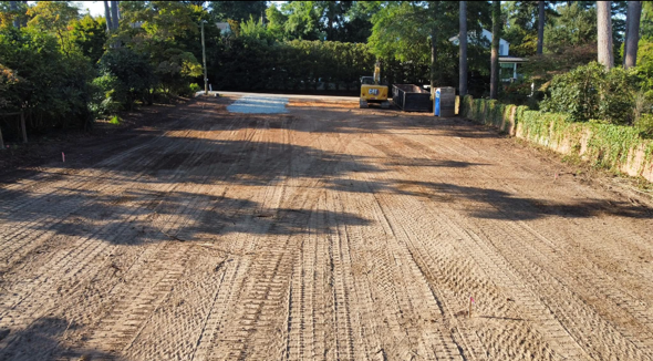 Graded dirt road under construction, bordered by trees and a hedge in sunlight