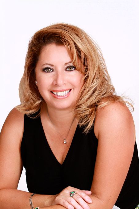 A smiling woman with shoulder-length highlighted hair, wearing a black top and jewelry, leaning forward against a backdrop.