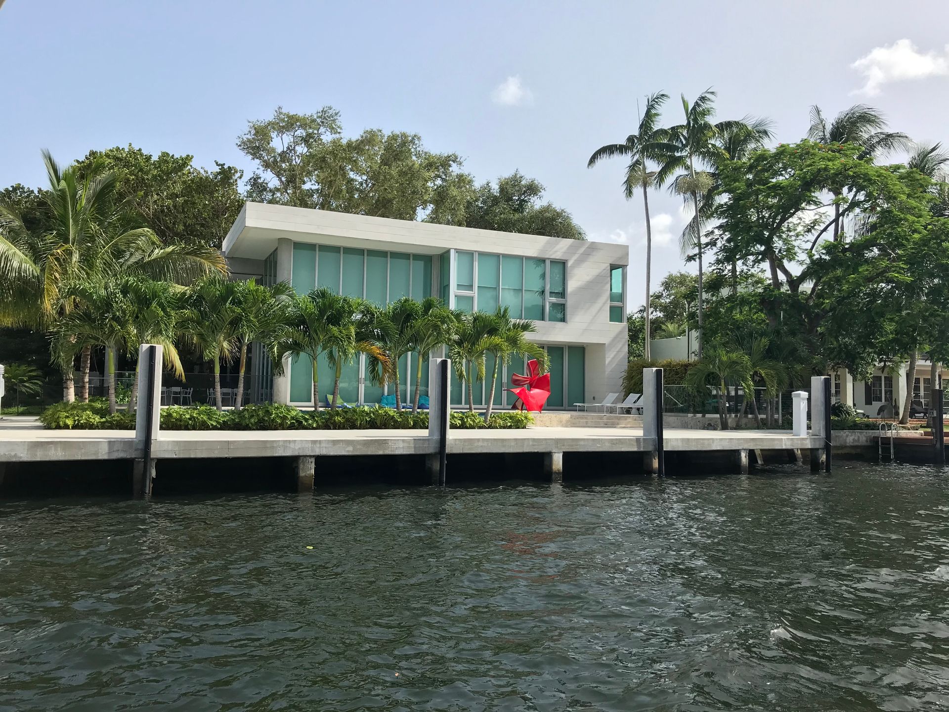 A modern white two-story house with large glass windows sits along a waterfront, flanked by palm trees and lush foliage.