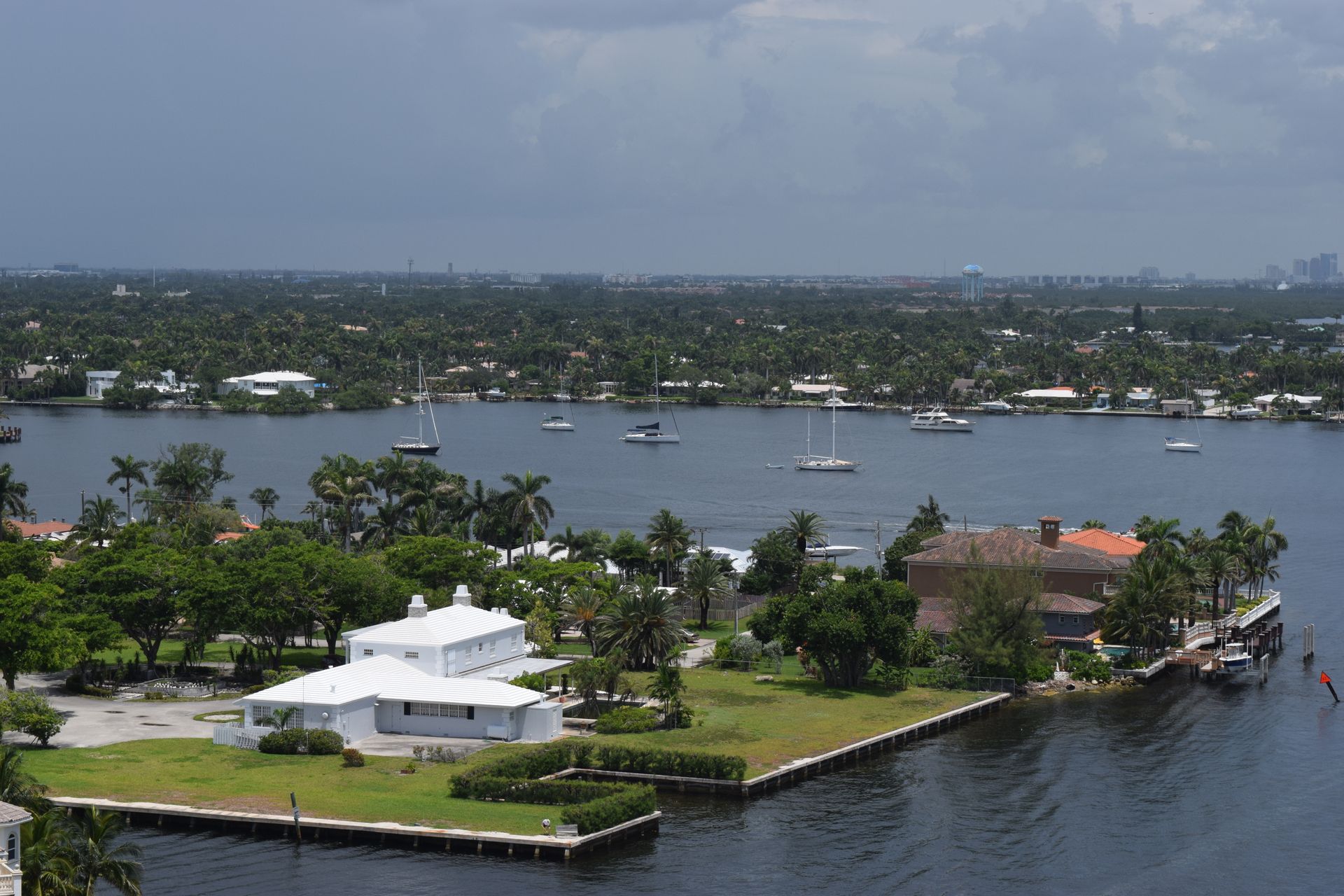 An aerial view of waterfront properties with white houses, lush green lawns, and boats on a calm bay under a cloudy sky.