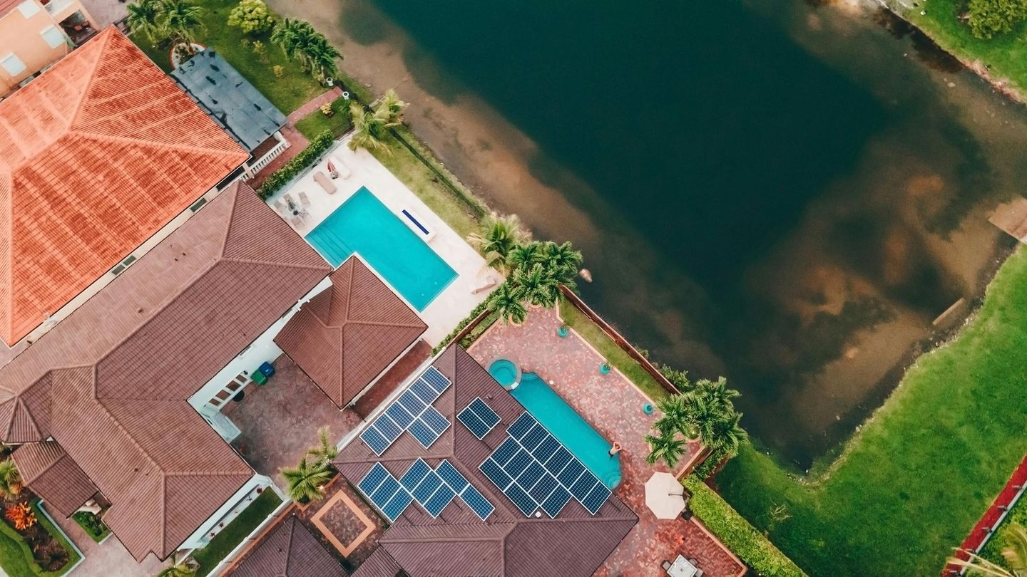 Aerial view of two adjacent residential homes with tile roofs, swimming pools, and solar panels, bordering a dark lake.