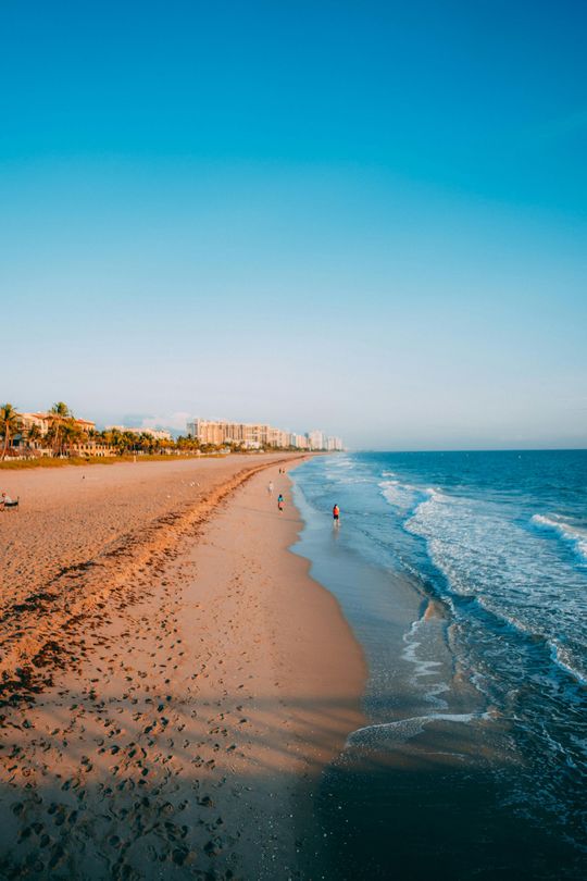 A wide, sunny beach with gentle waves meeting the shore, lined with distant hotels and palm trees under a clear blue sky.