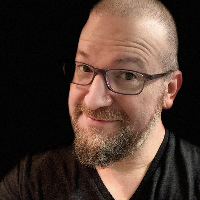 Photo of Drory Ben-Menachem — glasses, shaved head, beard, smiling, dark shirt, against black background.