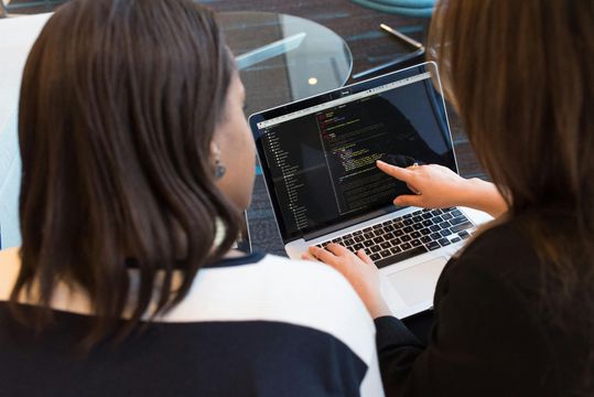 Two women are sitting at a table looking at a laptop computer.