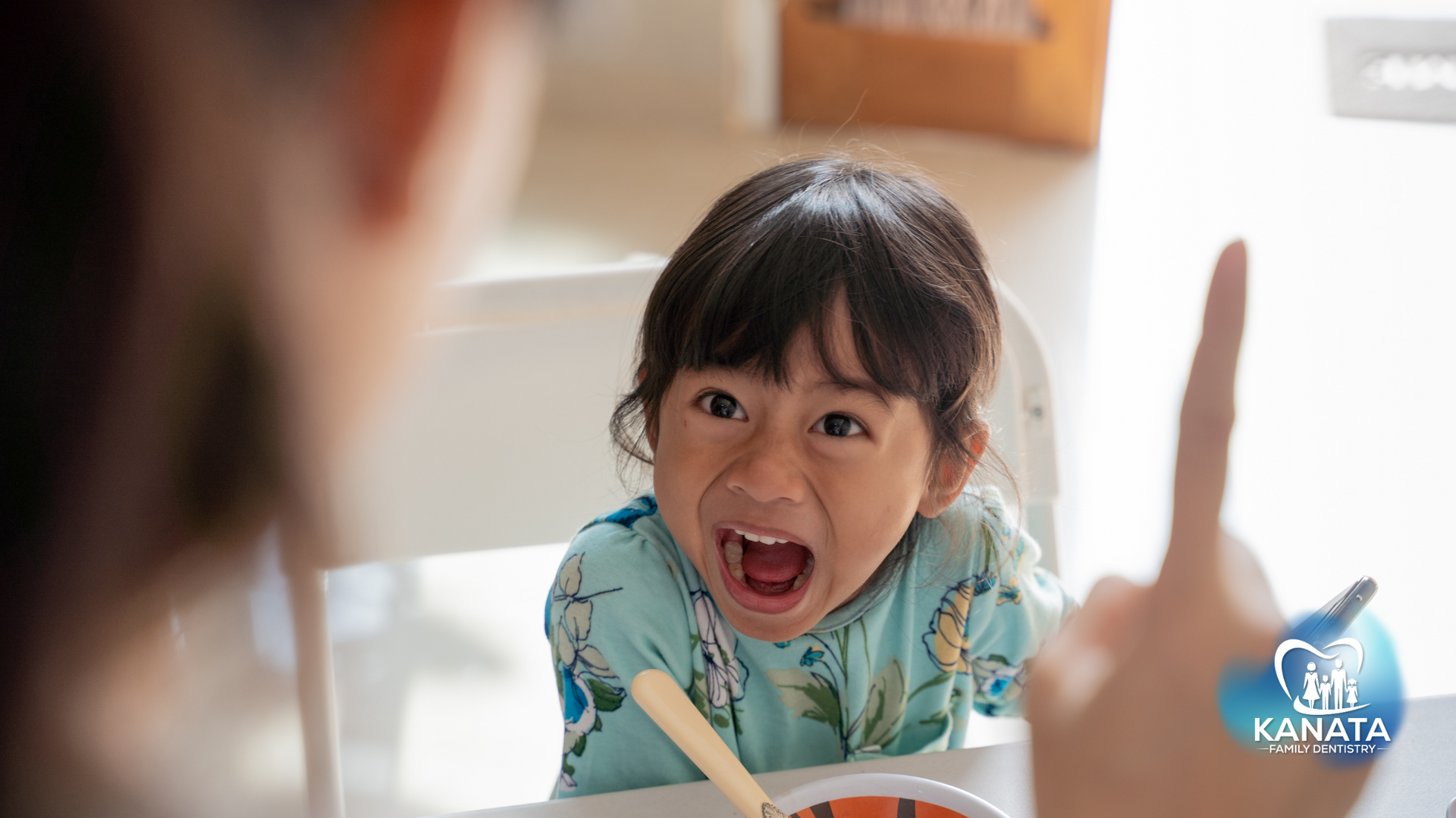 A little girl is sitting at a table with her mouth open and a woman is pointing at her.