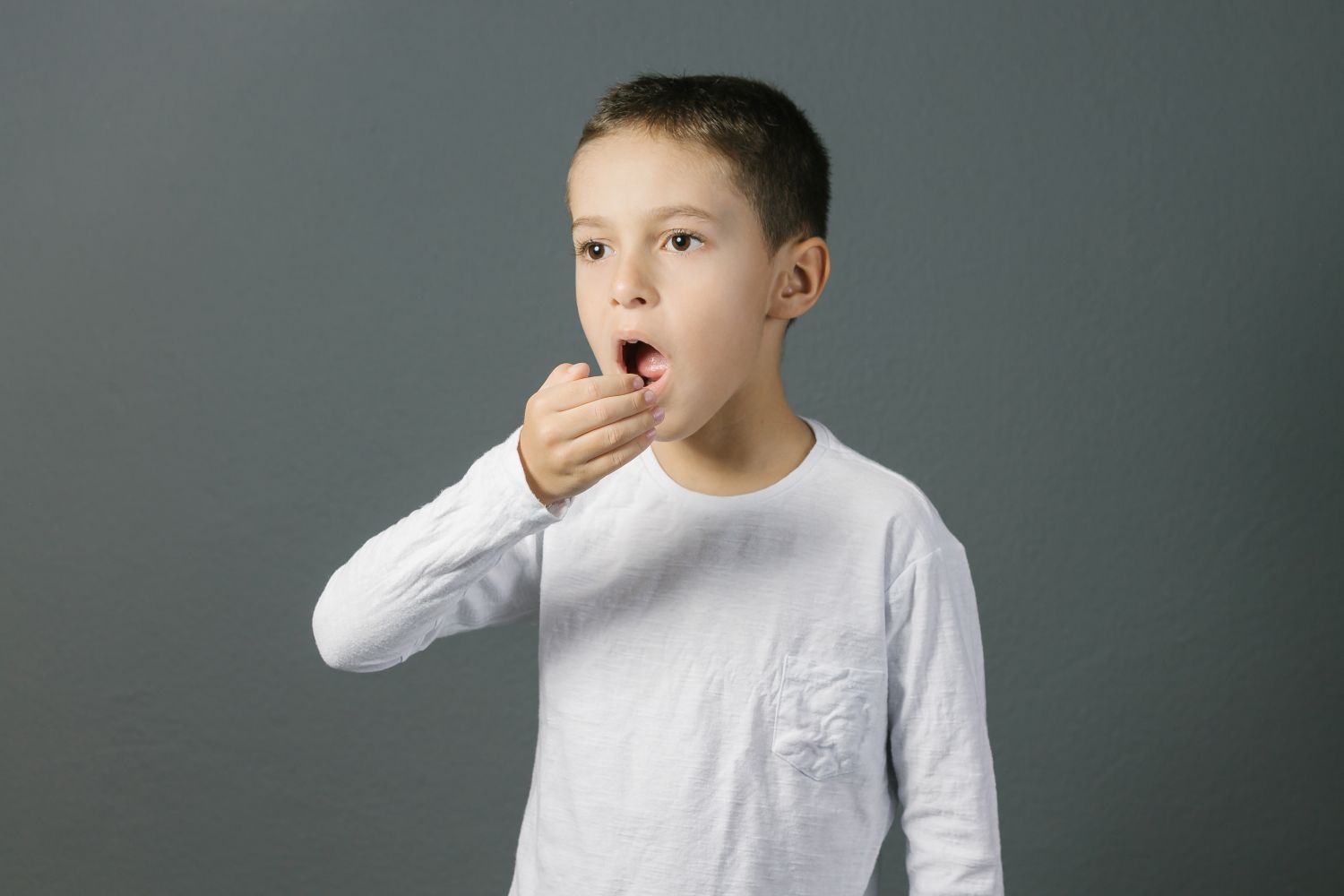 Boy with mouth open, hand raised to mouth, against a gray background.