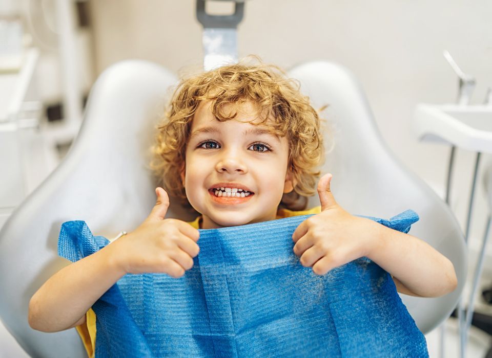 Boy with curly hair gives thumbs up in a dentist's chair, wearing a blue bib.
