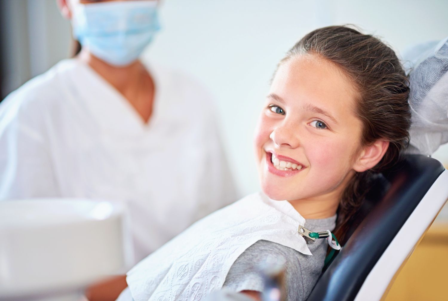 Girl smiles in a dentist's chair, dental hygienist in background.